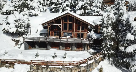 Cozy chalet with rustic wood architecture, large windows, snowy pine backdrop, stone terrace, and panoramic views of Auron's winter resort charm.
