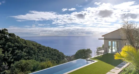 Sleek villa with infinity pool overlooking lush hills and azure sea, expansive windows, sun-soaked terrace, and manicured lawn.