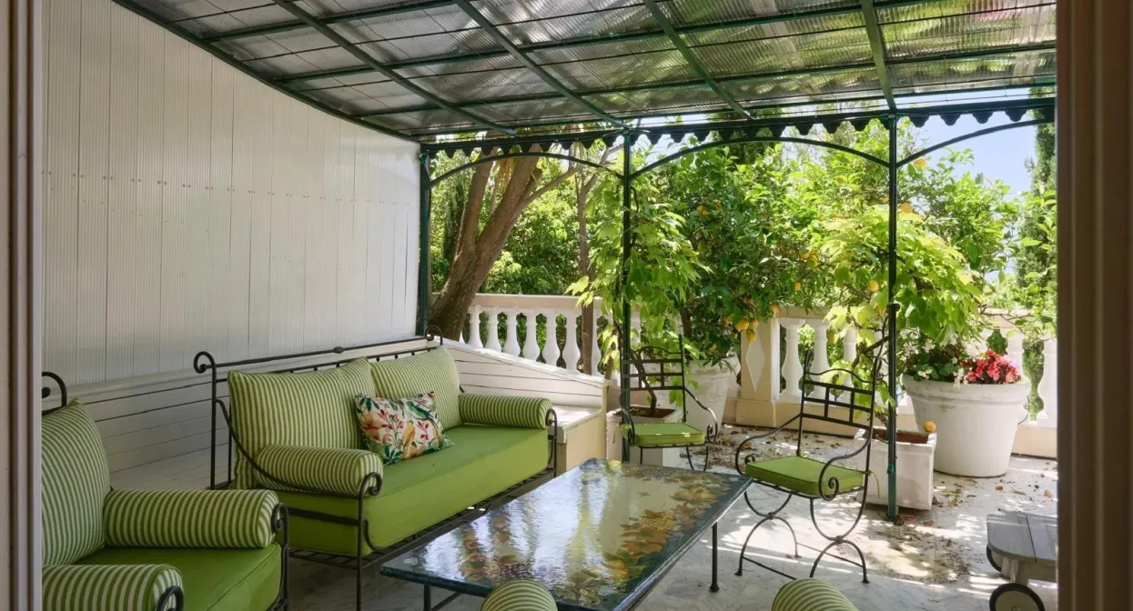 Covered terrace with elegant green seating, glass table, lush garden views, ornate iron details, and natural stone flooring, exuding Mediterranean charm.