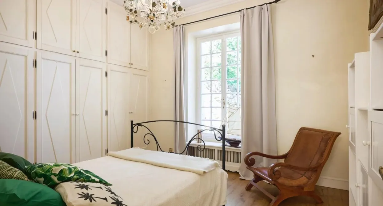 Elegant bedroom with vintage chandelier, wrought iron bed, classic white wardrobes, large window with curtains, and warm wooden flooring.