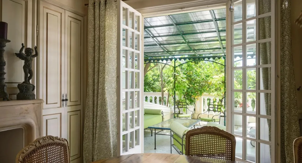 Elegant dining room opens to lush covered terrace with ornate ironwork and verdant views, featuring classic wood cabinetry and refined furnishings.