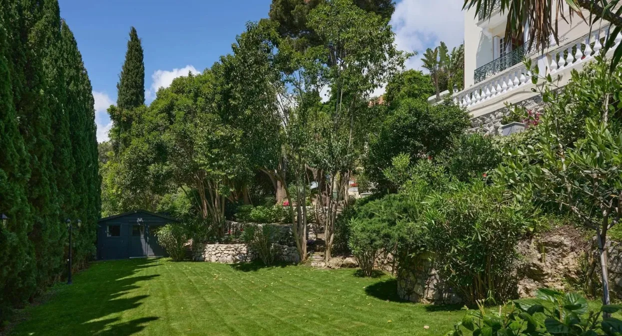 Lush garden with manicured lawn, mature trees, and stone accents, nestled beside a luxurious villa near Monaco's border.