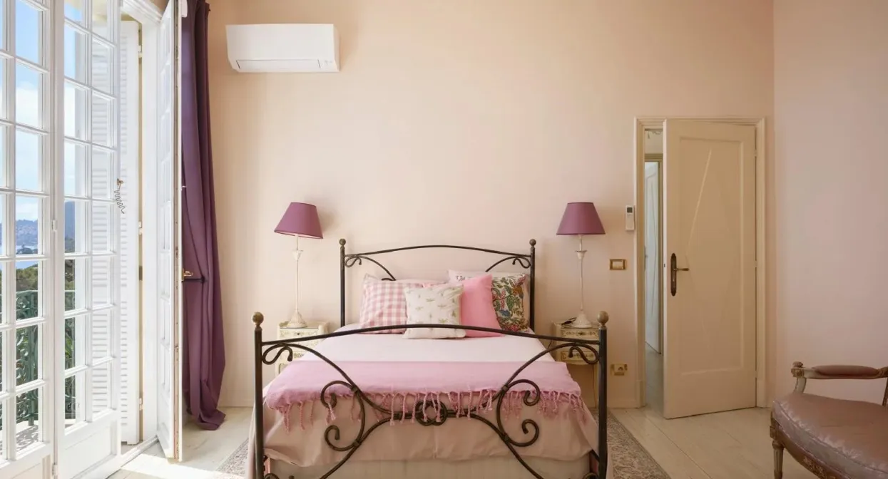 Elegant bedroom with ornate metal bed, soft pink accents, tall French doors opening to a balcony, and a chandelier, creating a serene, airy ambiance.