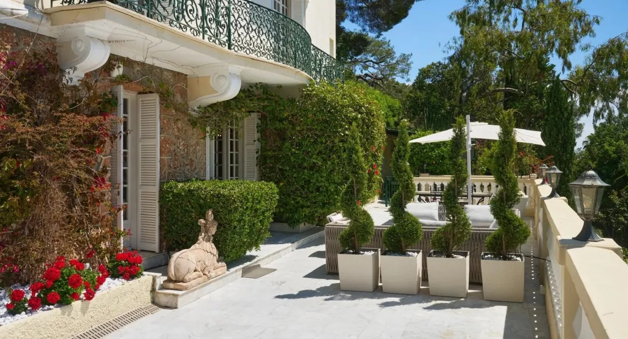 Elegant villa terrace with ornate iron railings, lush greenery, and sculptures; spacious marble flooring offers serene garden views.