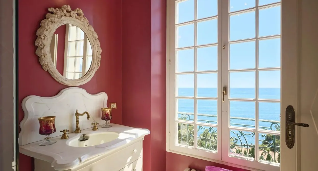 Luxurious villa bathroom with pink walls, ornate mirror, elegant white vanity, large window, and stunning Mediterranean Sea views.