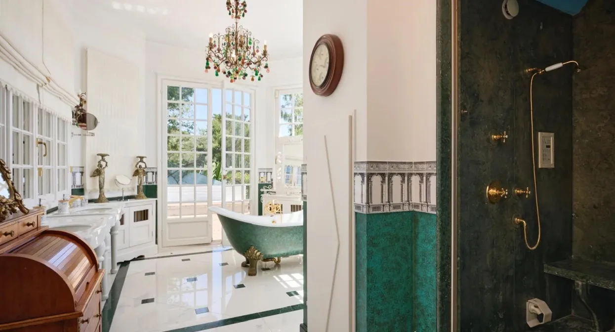 Elegant bathroom with freestanding green tub, chandelier, and ornate fixtures; sunlit via large windows with lush garden views near Monaco.